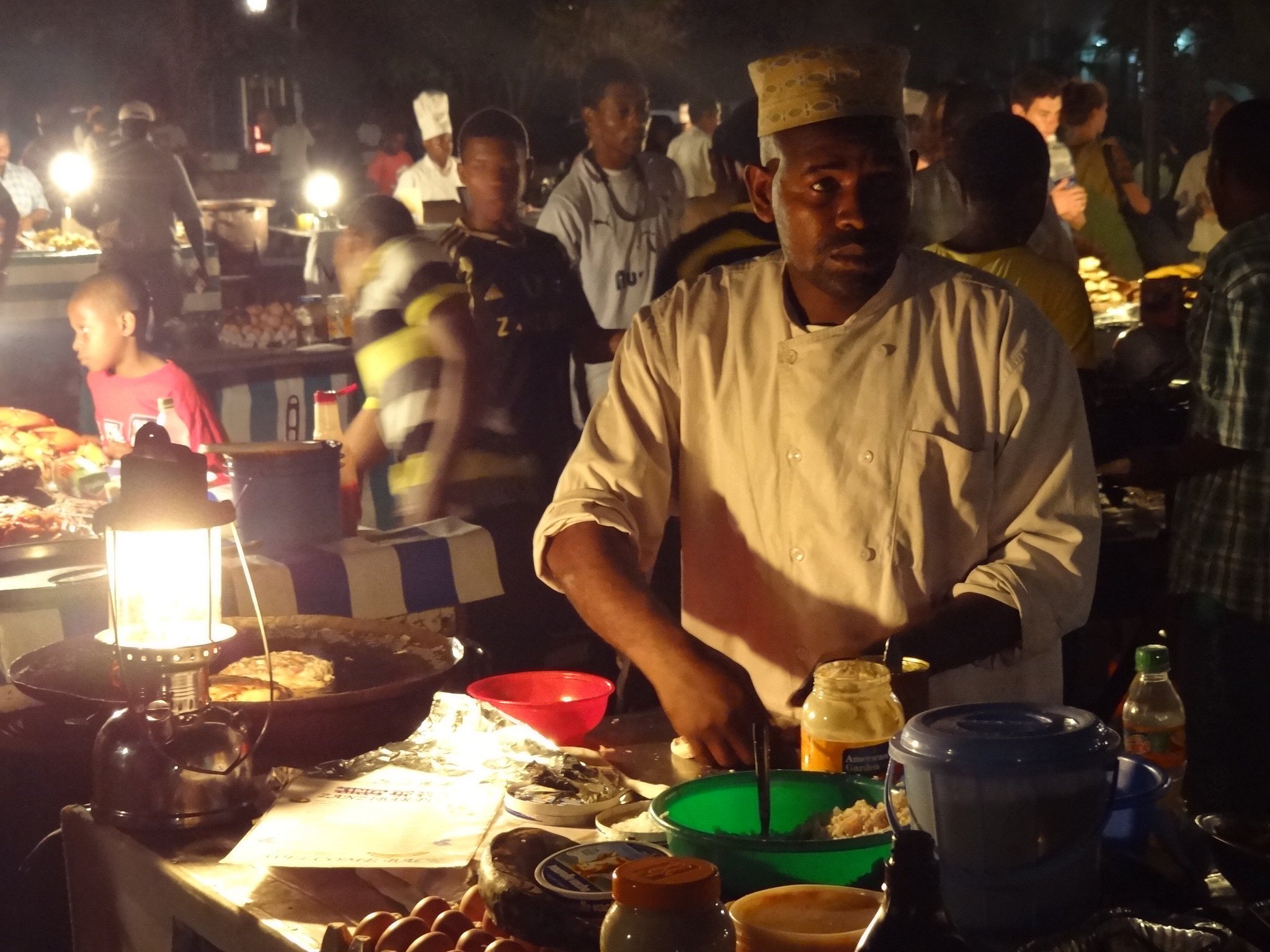 A food vendor in a white jacket and embroidered cap cooks under lantern light at the Forodhani Gardens night market in Stone Town, Zanzibar