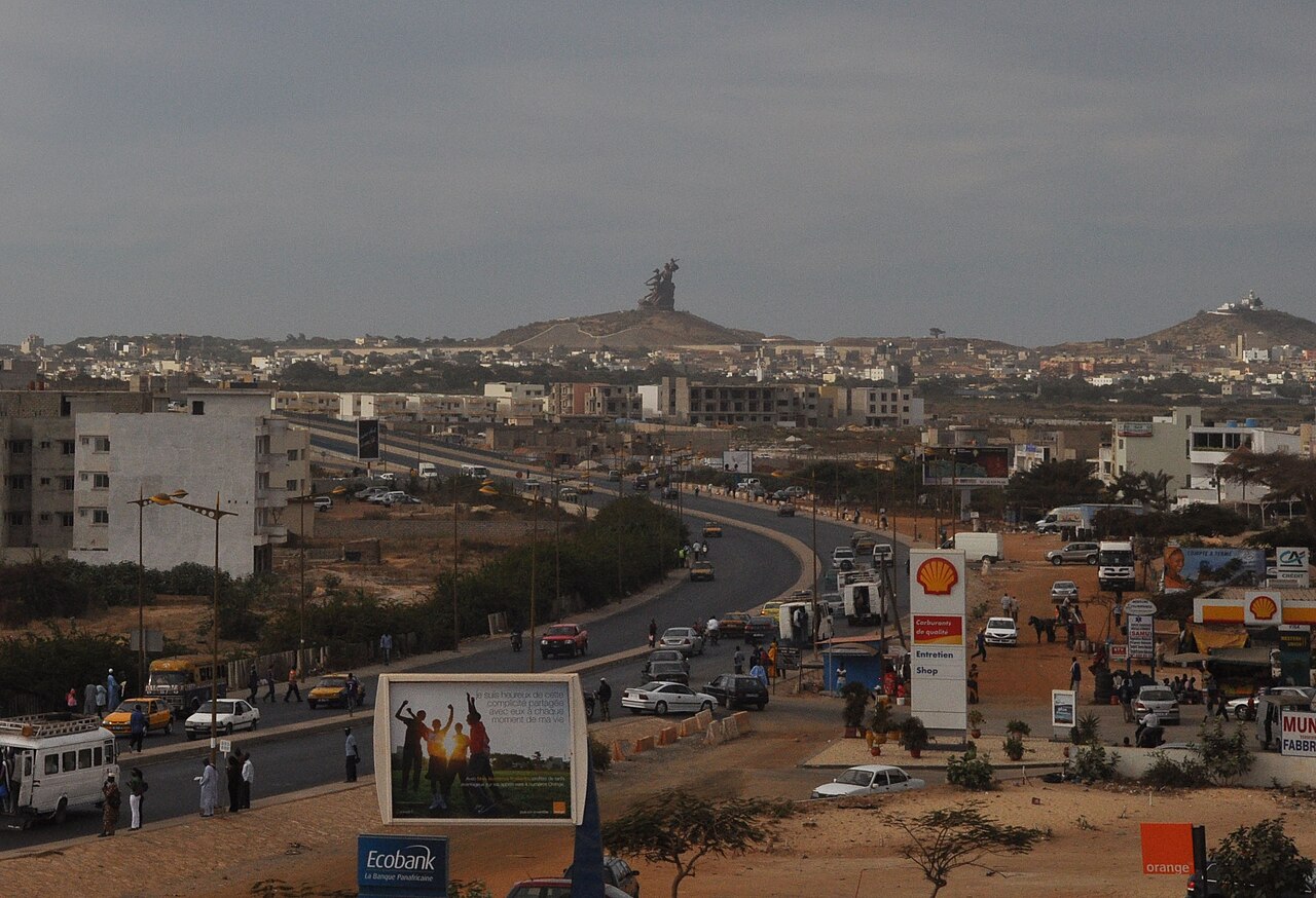 A busy road in Dakar with minibuses, taxis and billboards, and the African Renaissance Monument on a hill in the distance