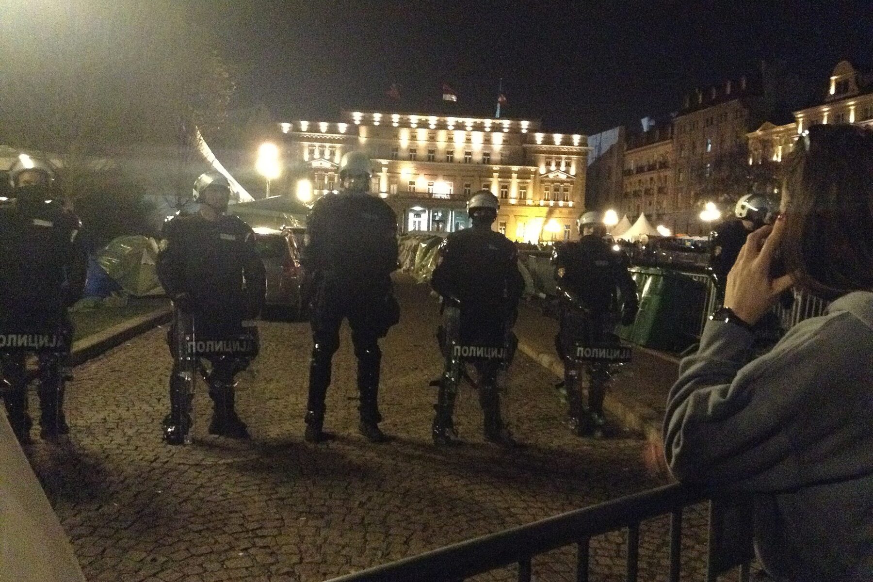Riot police stand in a line in Belgrade at night in front of a lit government building