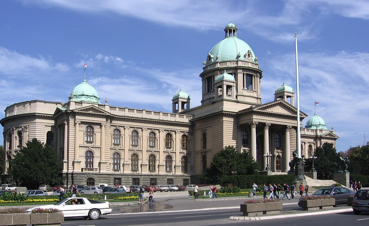 The House of the National Assembly of Serbia in Belgrade, a large neoclassical building with columns and green copper domes, seen from the street