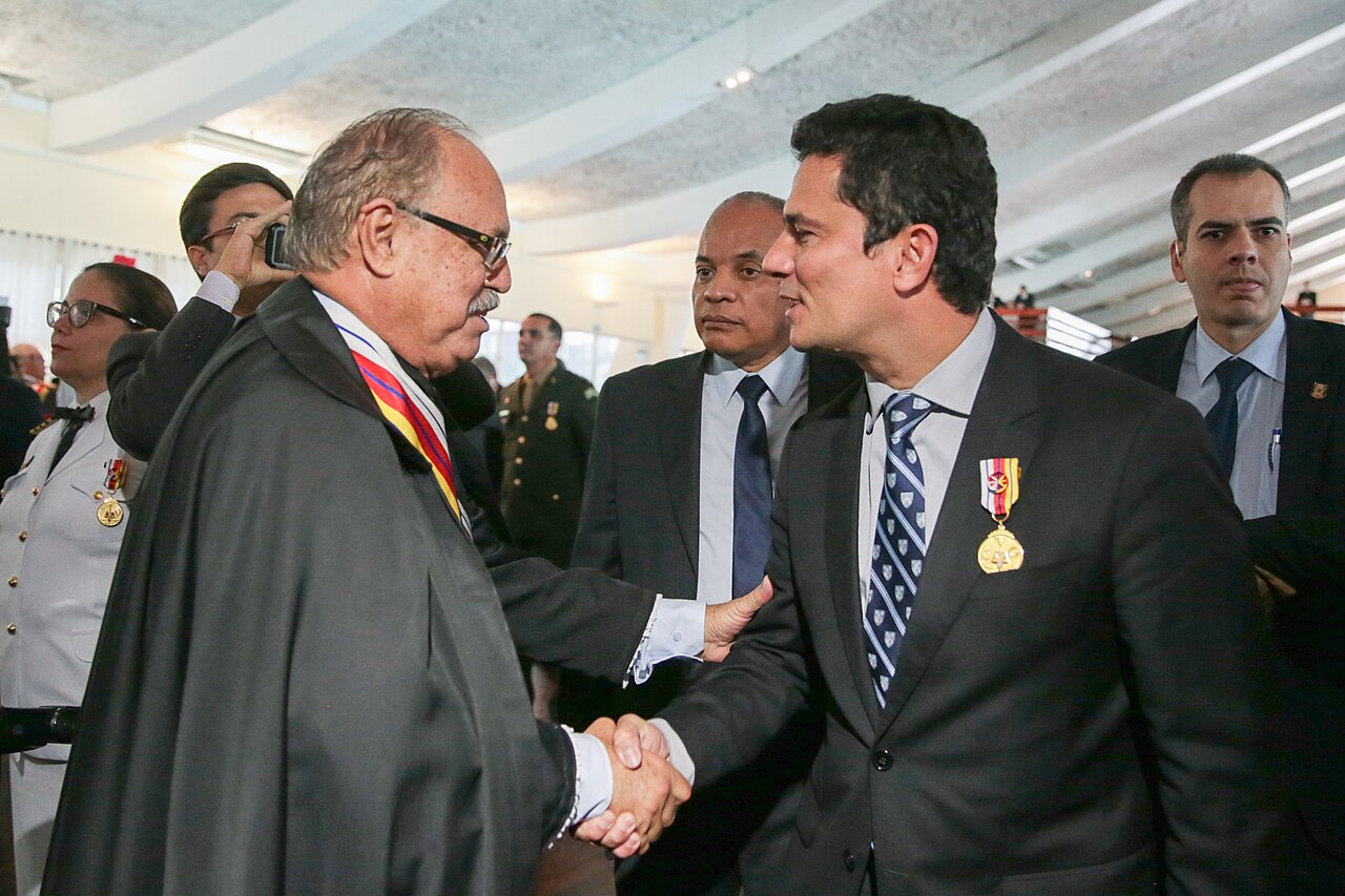 Sergio Moro, wearing a medal on his suit jacket, shakes hands with an older man in a black robe at a ceremony
