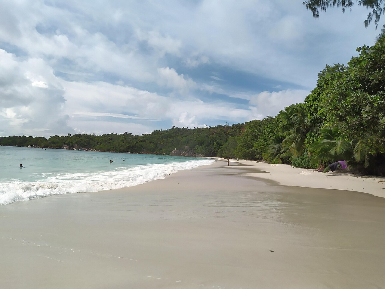 Waves roll onto the pale sand of Anse Lazio beach on Praslin, Seychelles, with swimmers and green forest on the headland