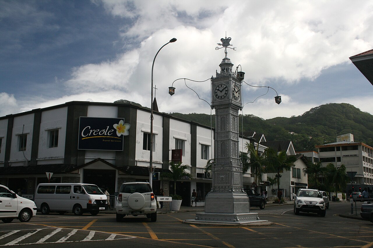 The silver clock tower at a street junction in Victoria, Seychelles, with shops, parked cars and green hills behind