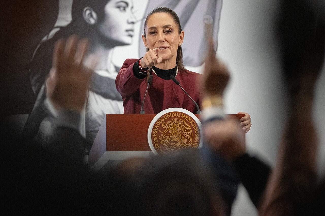 Mexican President Claudia Sheinbaum pointing to a reporter at the podium of a morning press conference, with raised hands in the foreground