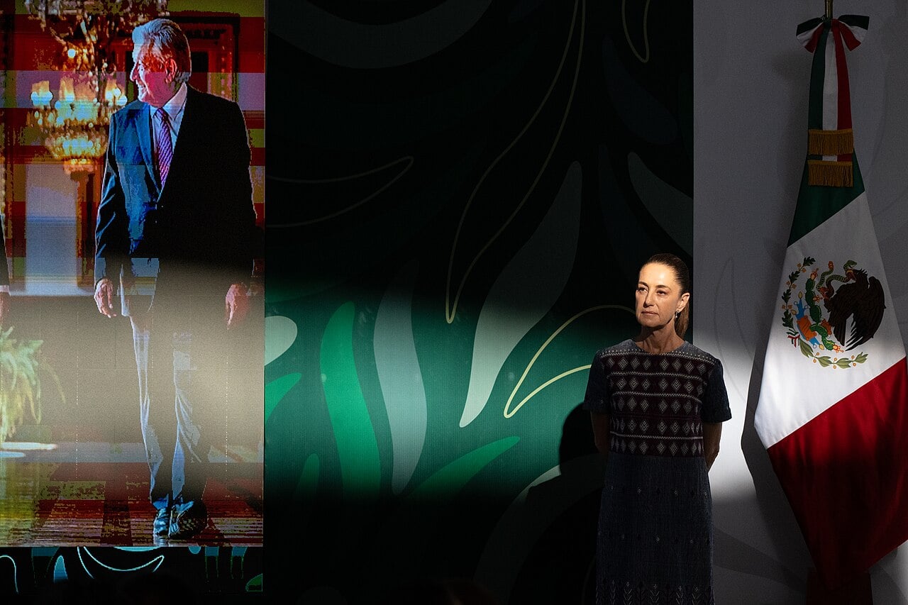 Mexican President Claudia Sheinbaum standing beside the Mexican flag at a morning press conference, with a video screen to her left