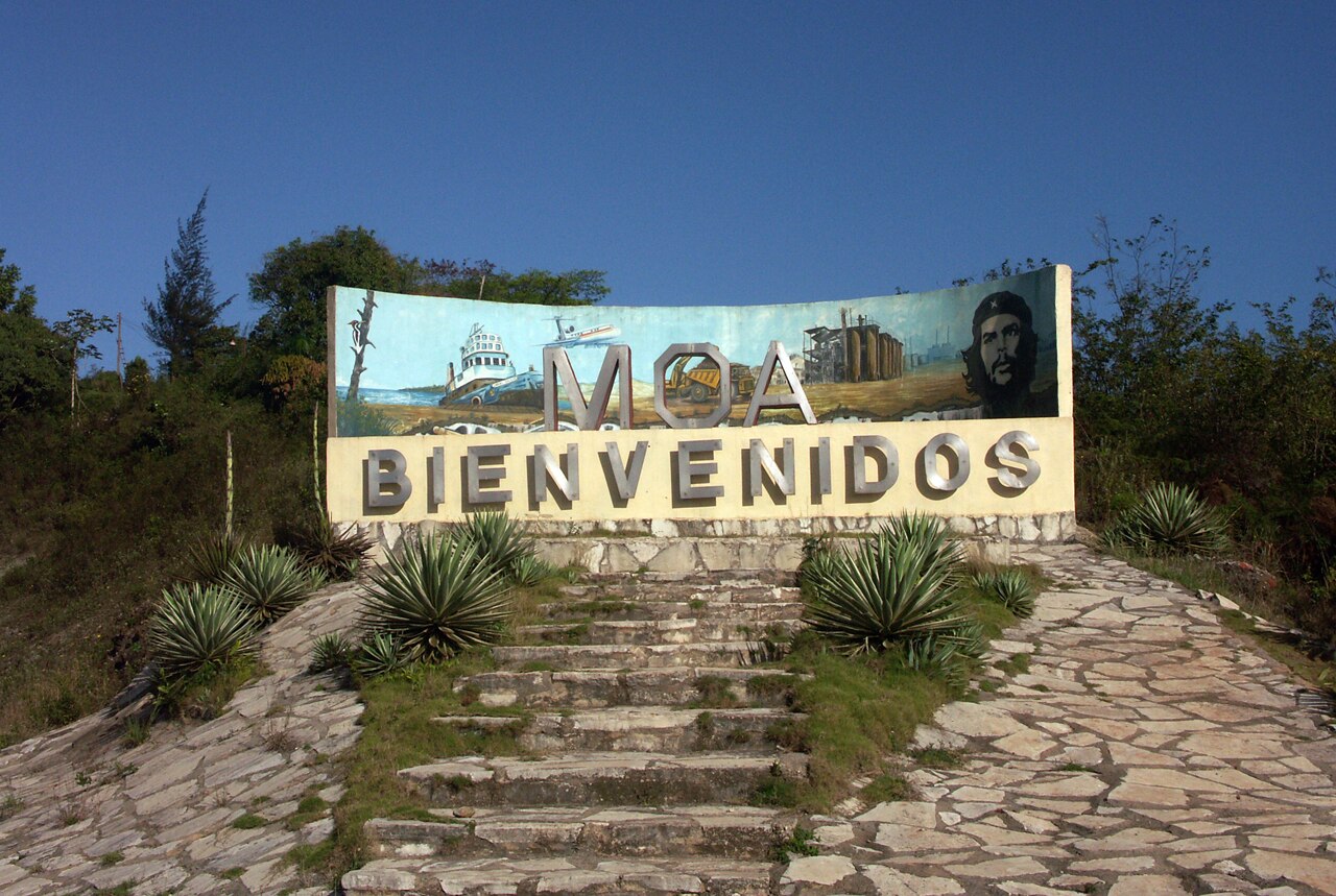 Welcome to Moa sign with a mural of a ship, a mining truck and a processing plant at the entrance to Moa, Cuba