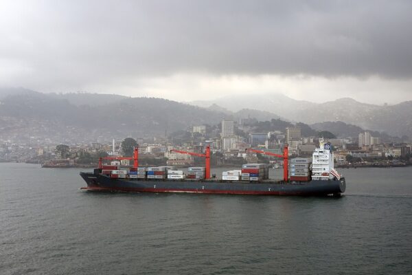 A container ship carrying Maersk boxes passes the hills and buildings of Freetown, Sierra Leone, under heavy grey cloud