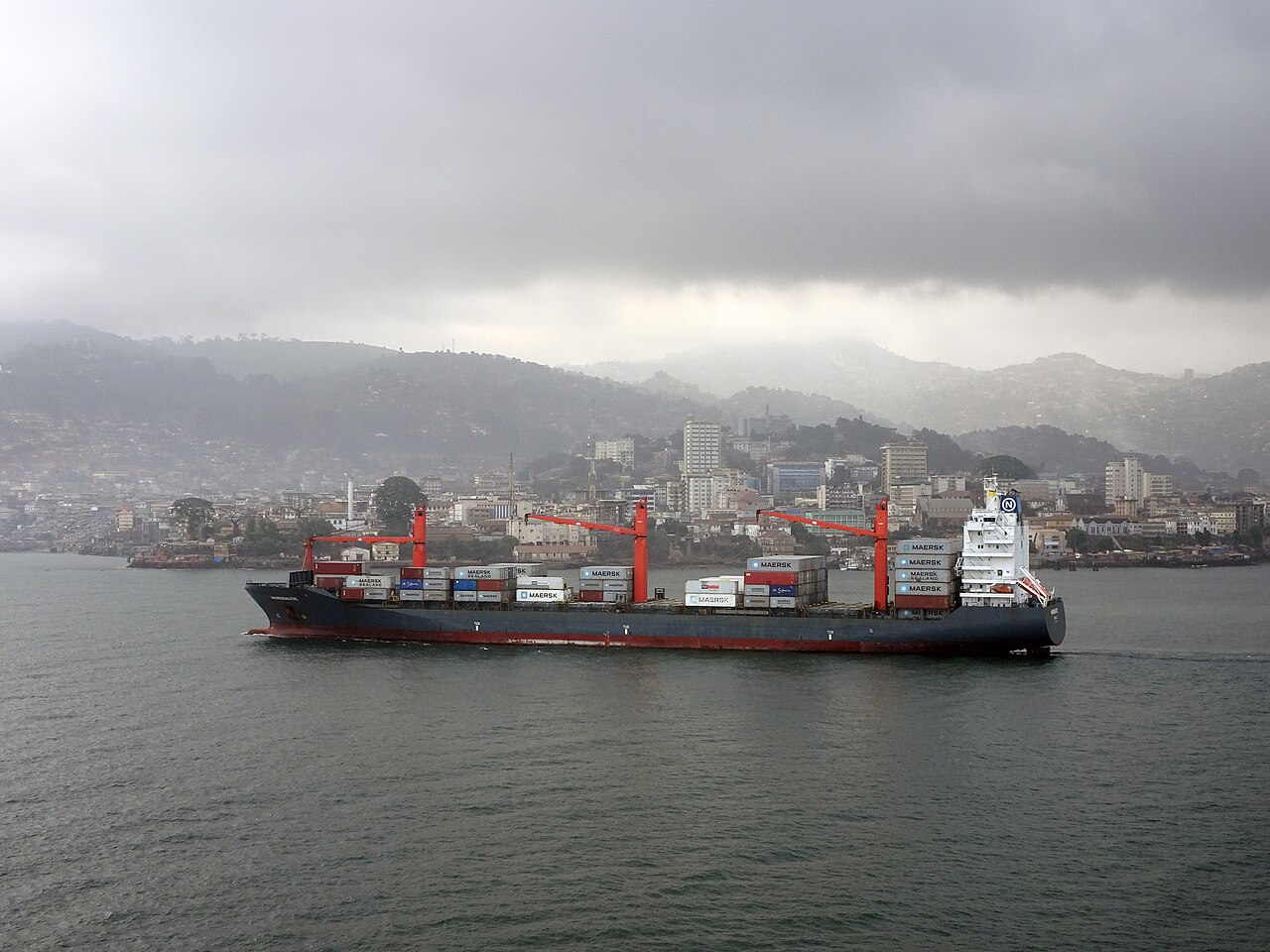 A container ship carrying Maersk boxes passes the hills and buildings of Freetown, Sierra Leone, under heavy grey cloud