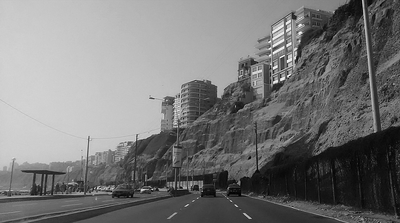 Black-and-white view of the Costa Verde coast road in Lima, with cars driving below steep cliffs topped by apartment towers and a bus shelter on the left.