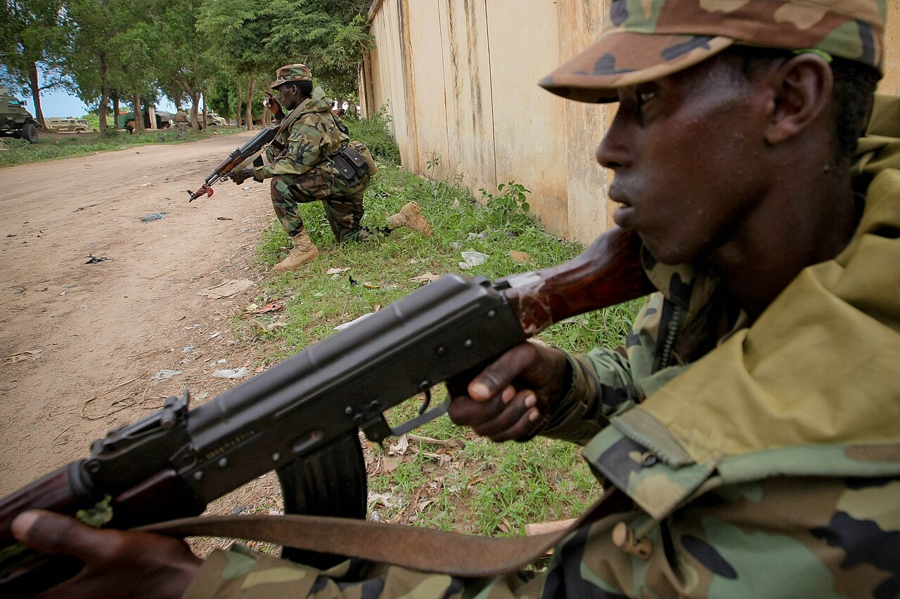 Two Somali soldiers in camouflage take defensive positions beside a wall on a dirt road in Afgooye