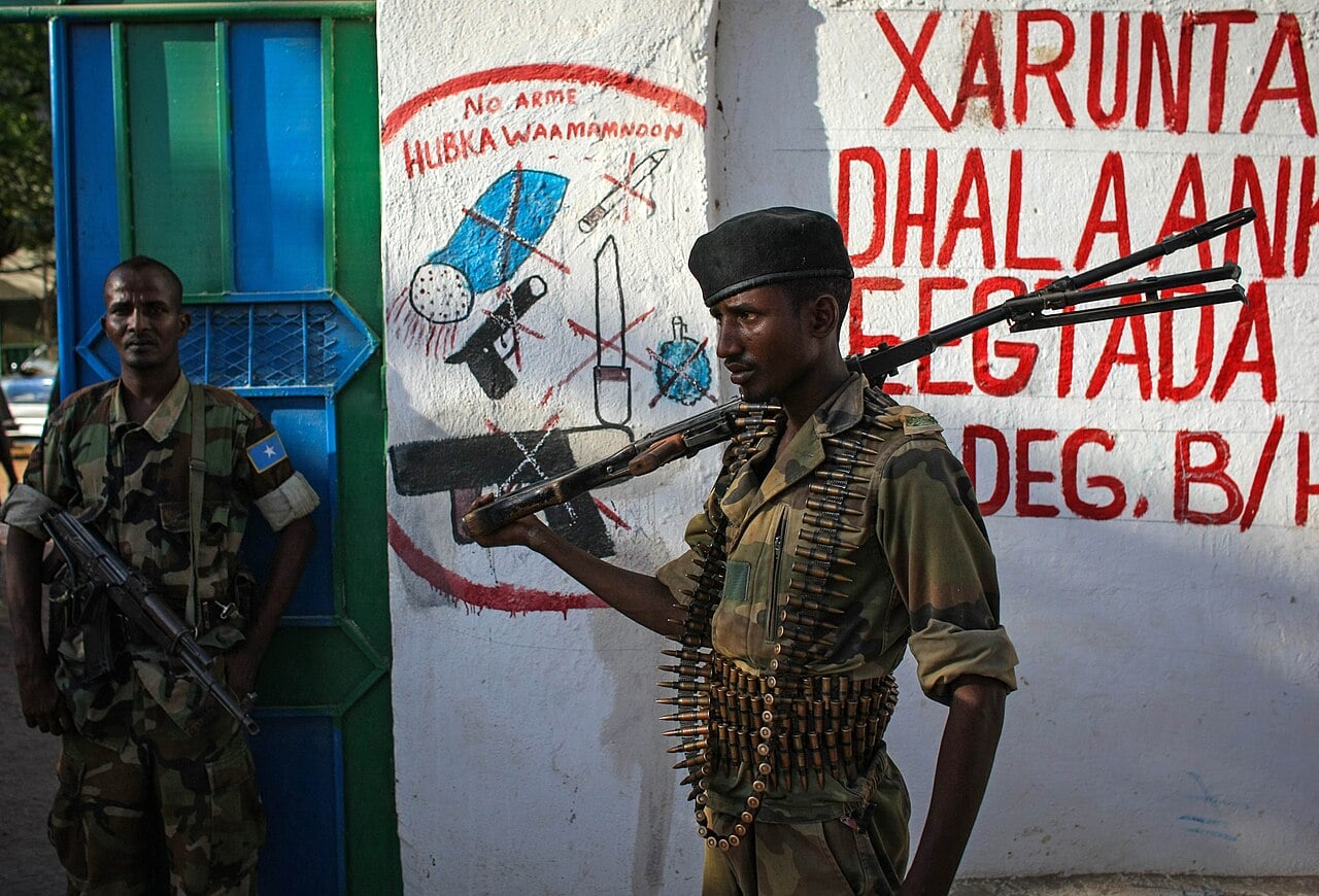 Two Somali National Army soldiers in camouflage stand guard beside a painted wall in Burhakaba, one carrying a machine gun with an ammunition belt