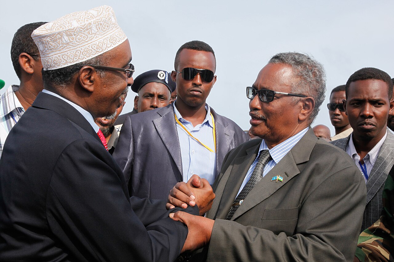 Two senior Somali officials in suits shake hands outdoors, surrounded by aides and a police officer