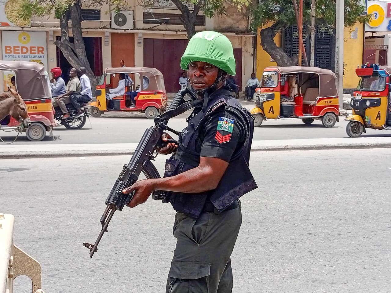 An African Union police officer with a rifle on a Mogadishu street, with yellow and red auto-rickshaws and a donkey cart behind him
