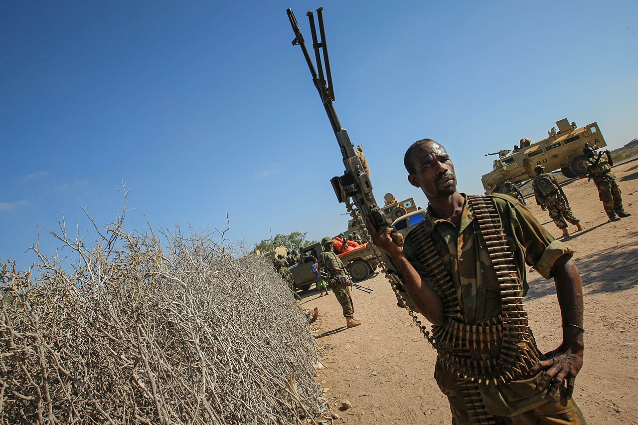 A Somali National Army soldier with a machine gun and ammunition belt stands in a dusty street while armoured vehicles and soldiers wait behind him