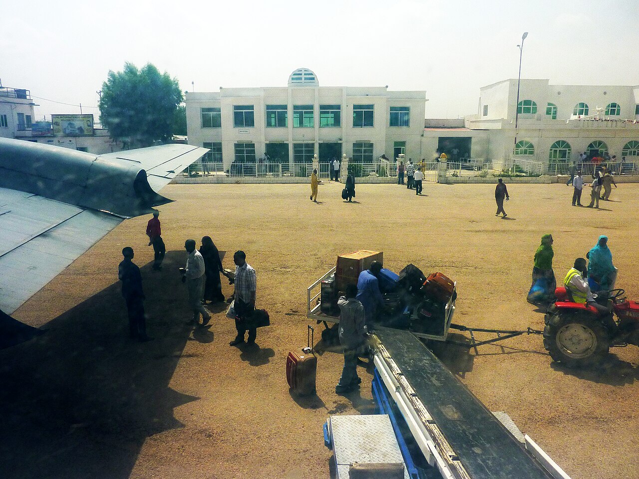 Passengers and baggage handlers on the apron at Hargeisa airport, with a white terminal building behind a tractor and trolley