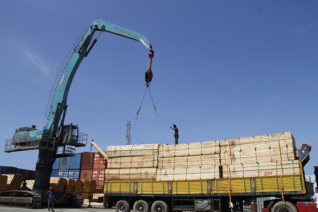 A crane lifts stacked timber onto a flatbed truck beside shipping containers at the Mogadishu seaport