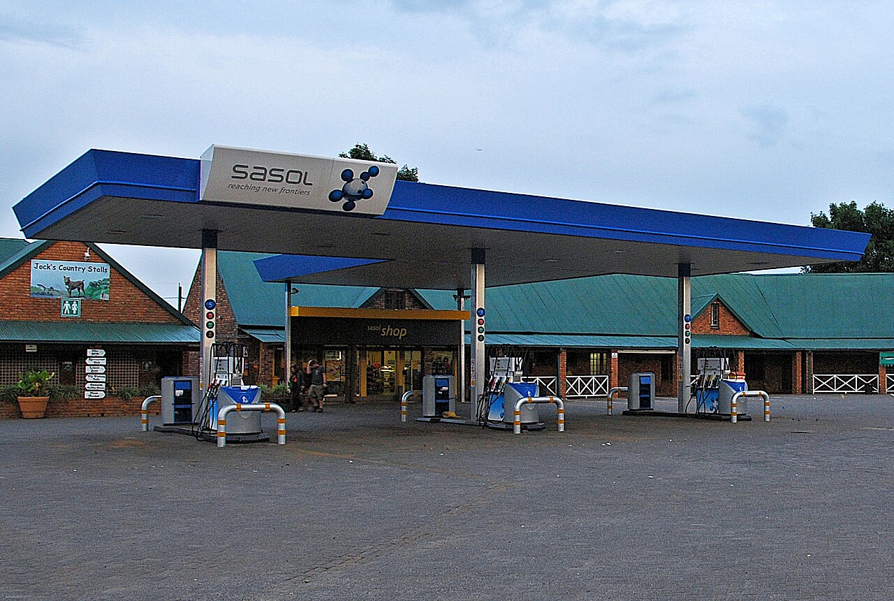 A Sasol petrol station with a blue canopy, fuel pumps and a small shop in front of brick buildings with green roofs, under a cloudy sky