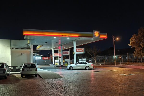A Shell petrol station in Plumstead, Cape Town, lit at night, with a white sedan parked beside the pumps and red V-Power signs under a yellow and red canopy