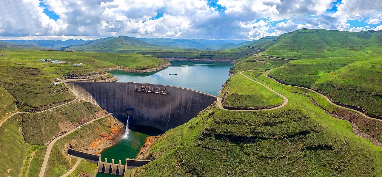 Katse Dam's curved concrete wall holding back a reservoir between green hills, with winding roads and a water release below the wall