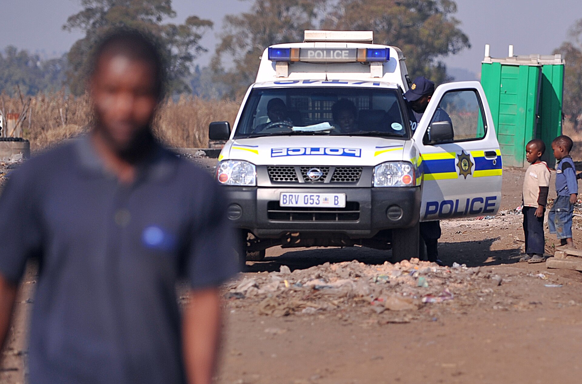 A South African Police Service van parked on a dirt road in an informal settlement near Johannesburg, with children watching