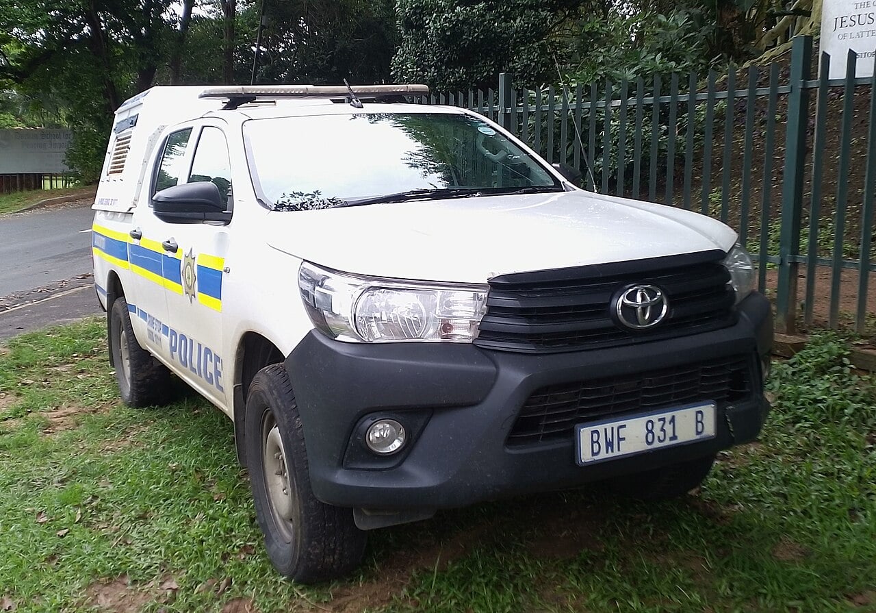 White South African Police Service Toyota Hilux pickup with blue and yellow stripes parked beside a fence