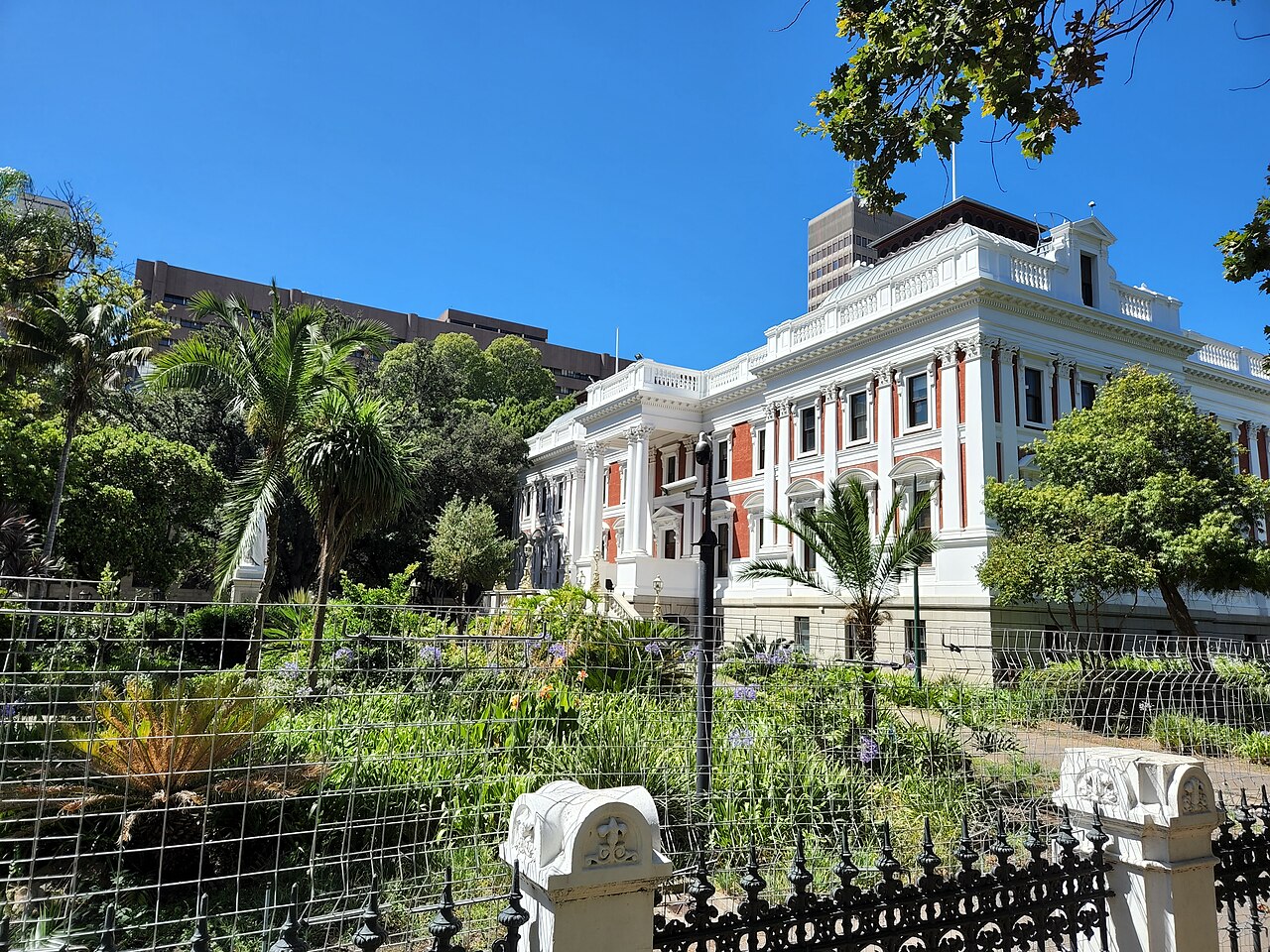 The white and red-brick Houses of Parliament in Cape Town seen through palms and garden greenery under a blue sky