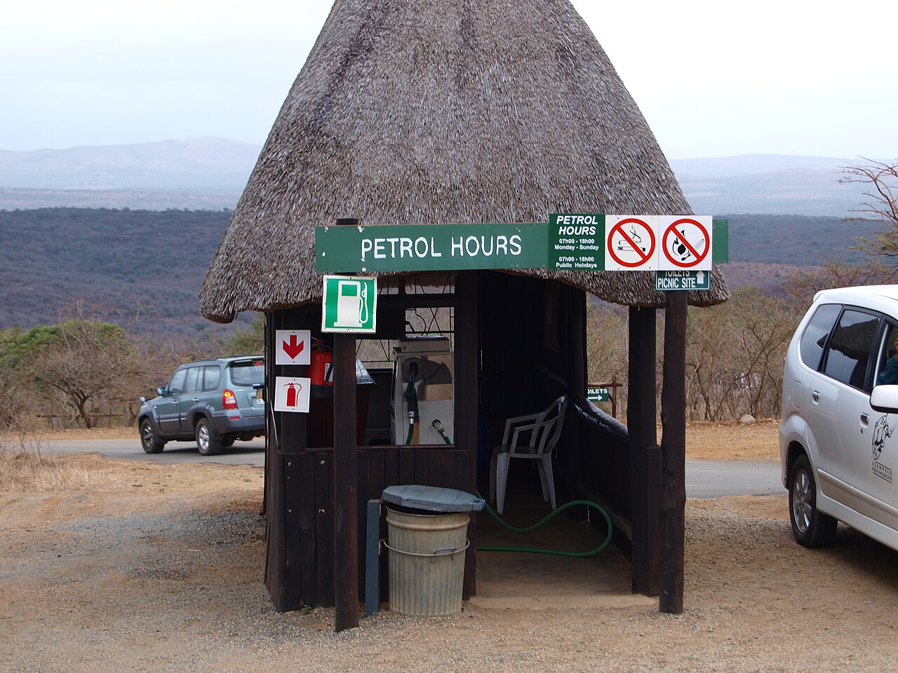 A fuel pump under a thatched roof with a Petrol Hours sign, next to parked cars and dry bush in South Africa
