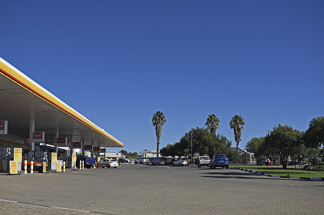 Shell service station canopy with V-Power pumps and parked cars under a clear blue sky beside palm trees