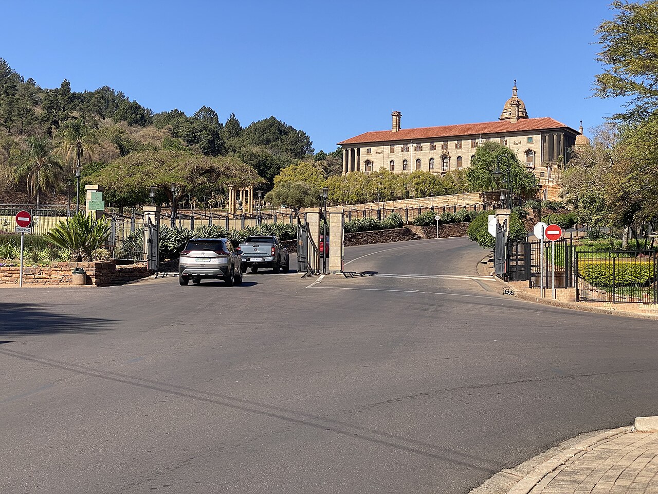 The sandstone Union Buildings in Pretoria with a red tiled roof and a dome, seen from a road and gate in the foreground under a clear blue sky
