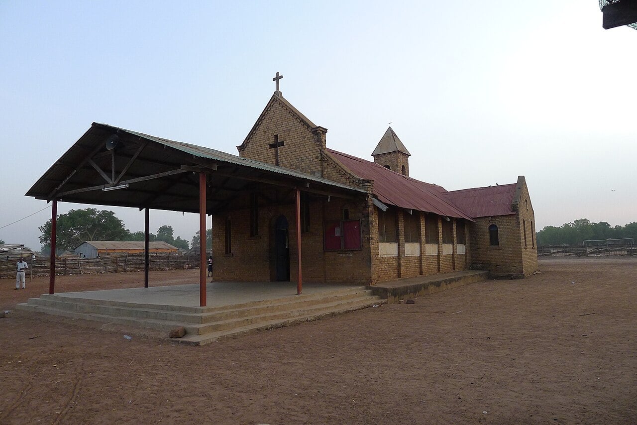 Brick Catholic church with a red metal roof and an open porch on bare red earth in Tonj, South Sudan