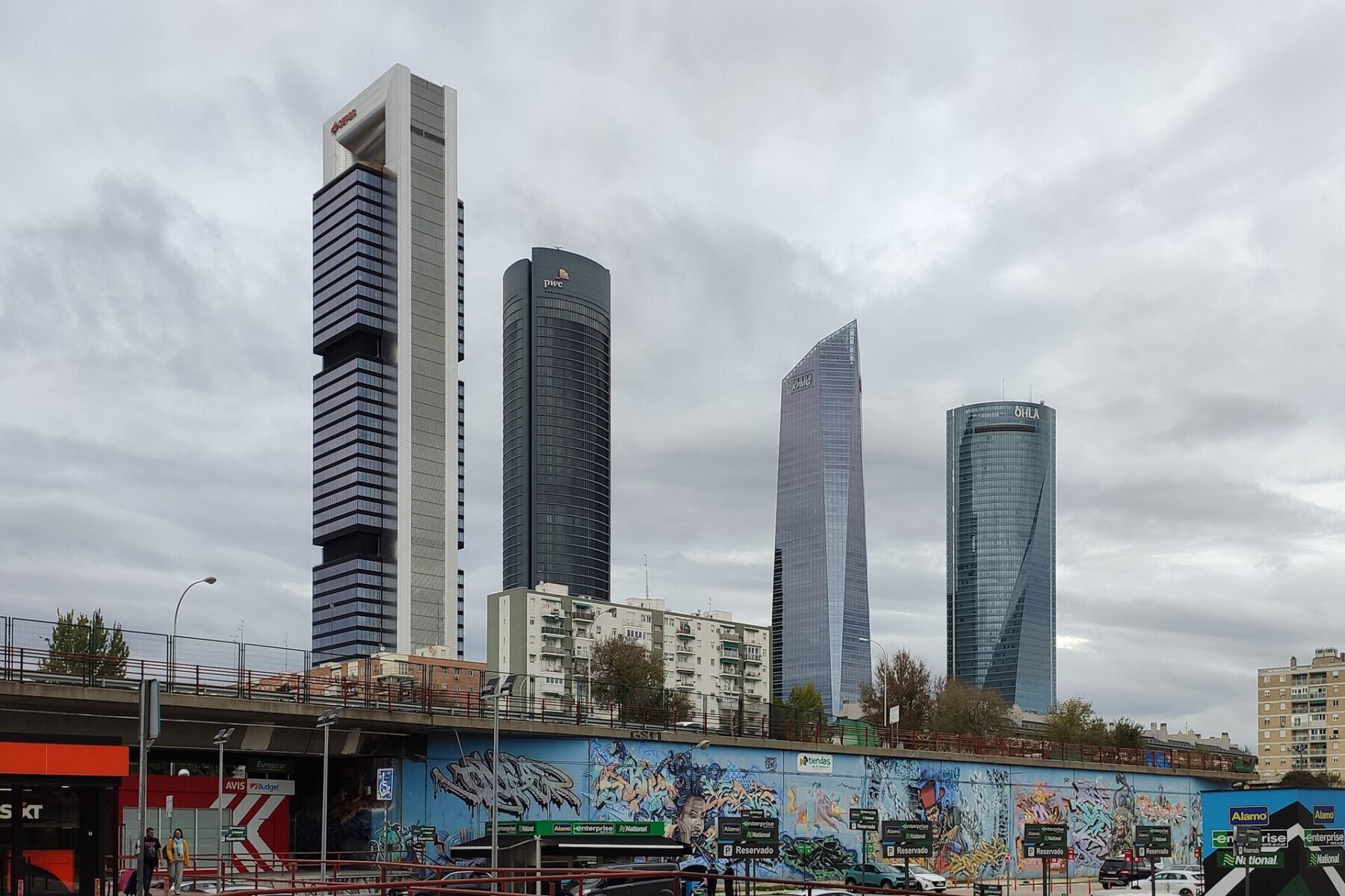 The four skyscrapers of Madrid's Cuatro Torres business district under a grey sky, with apartment blocks and a graffiti wall in front