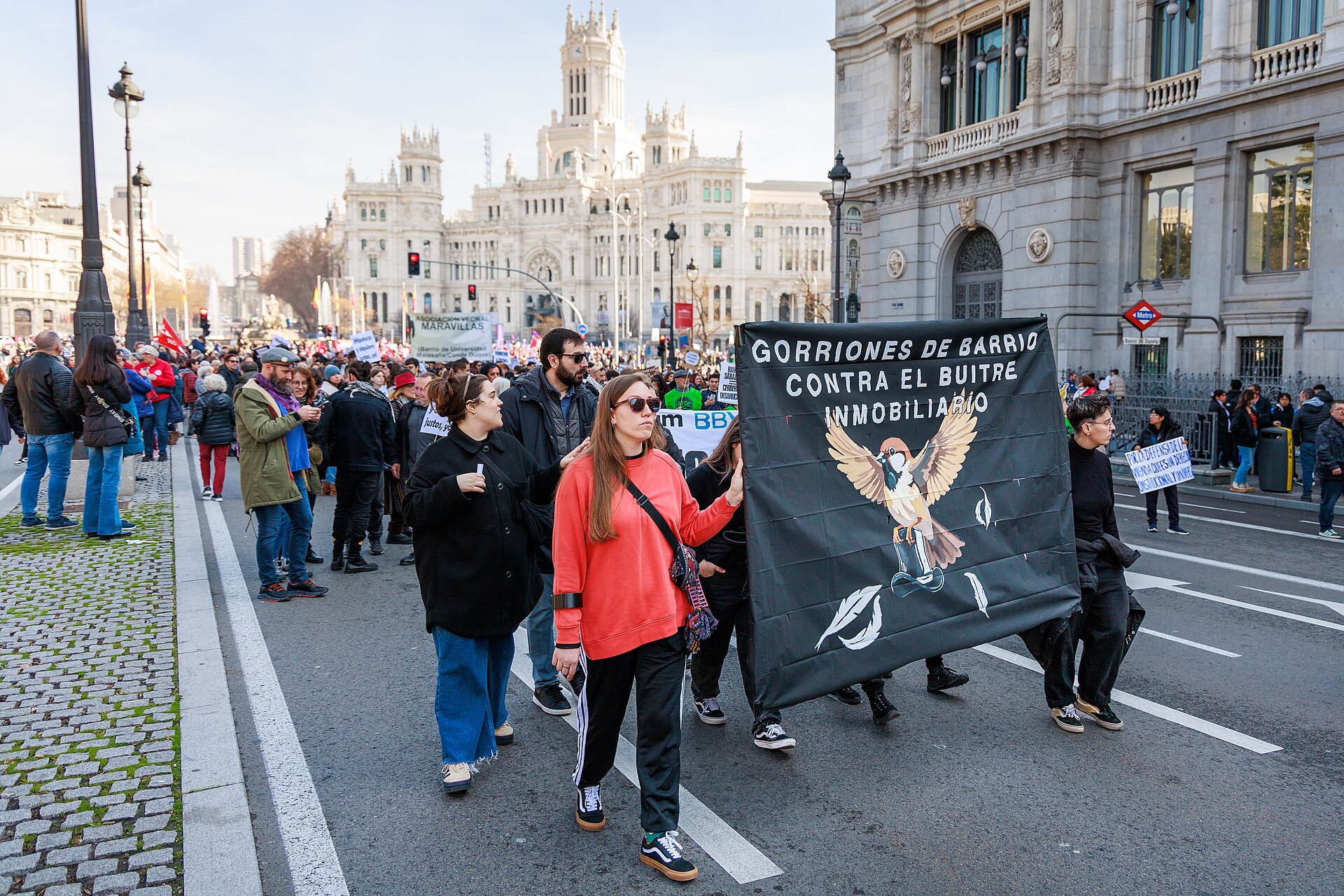 Protesters march down Calle de Alcalá in Madrid behind a black banner against property speculators, with Cibeles Palace behind them