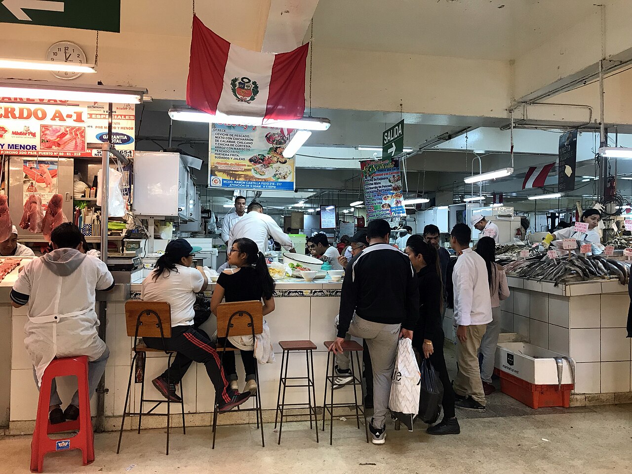 Diners on stools at a ceviche counter in Lima's Mercado Central, with a Peruvian flag and a green Salida exit sign overhead