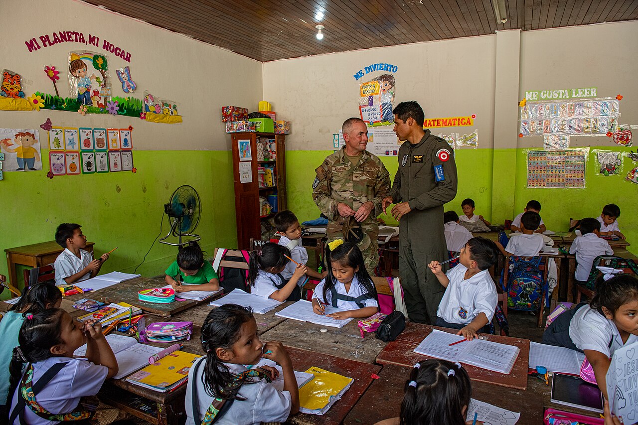 Children in white school uniforms write at wooden desks in a classroom in Iquitos, Peru, with Spanish signs on the walls and two air force officers behind them