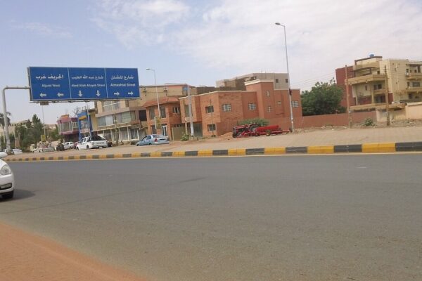 A wide road in Khartoum with a blue Arabic and English road sign, shops and houses, a white car at left