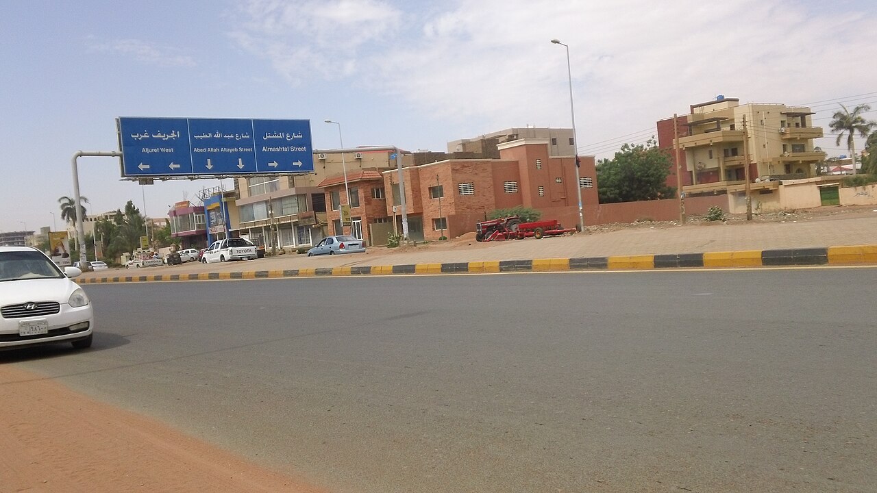 A wide road in Khartoum with a blue Arabic and English road sign, shops and houses, a white car at left