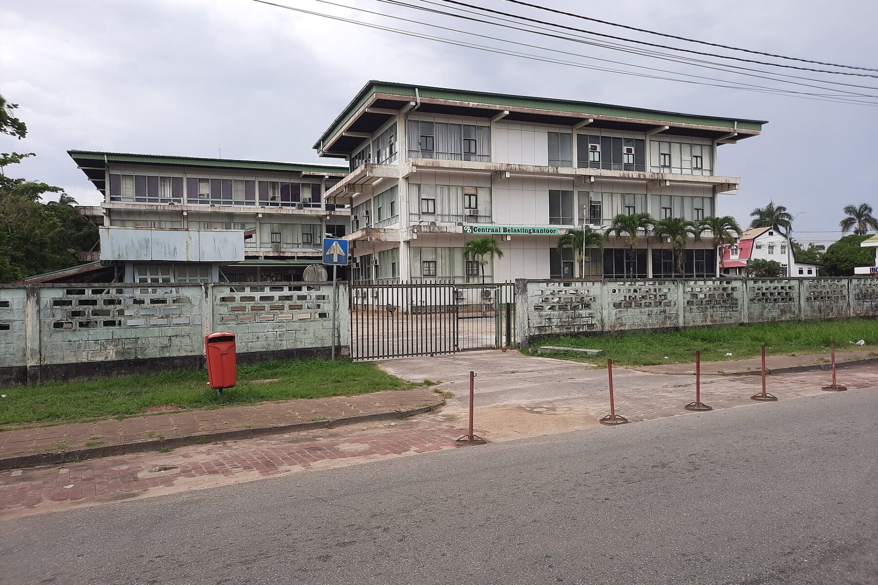 The three-storey Centraal Belastingkantoor, Suriname's central tax office, behind a gated wall on a street in Paramaribo under a grey sky