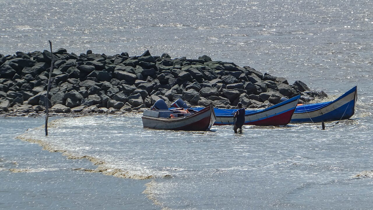 Three small wooden boats with outboard motors beside a rock breakwater on the Corantijn River, with a man standing in the shallow water