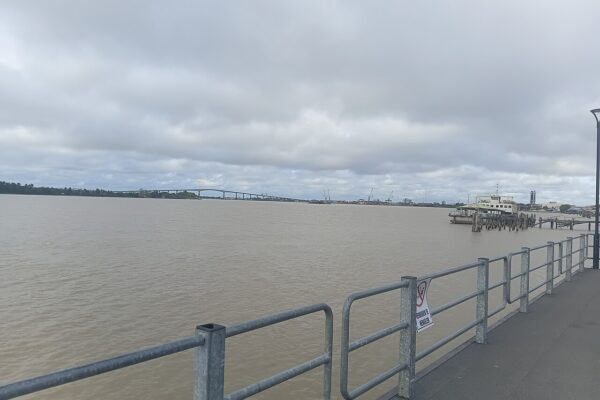 Wide brown river at Paramaribo seen from a railed pier, with a long road bridge and port cranes on the far shore under heavy cloud.