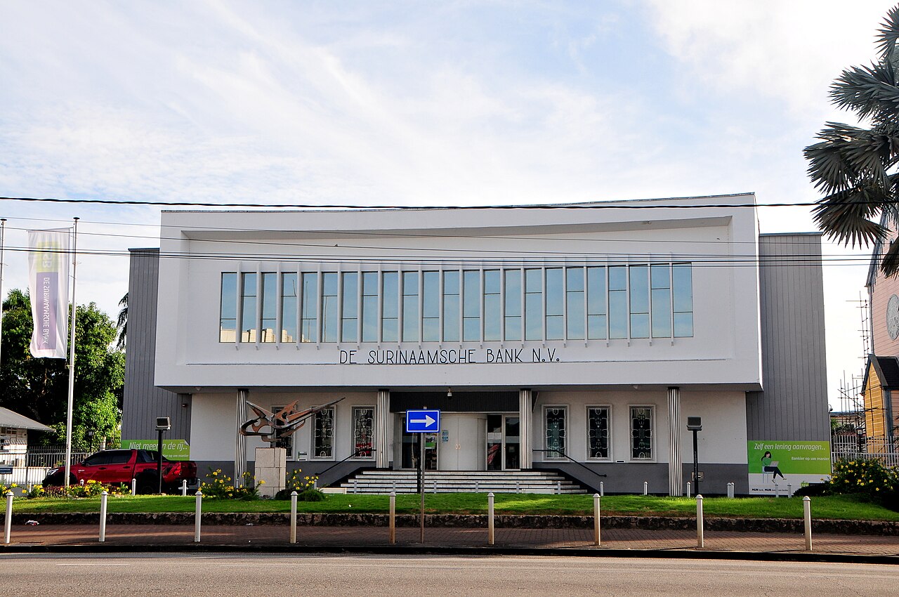 White modernist bank building with a long window band and the words De Surinaamsche Bank N.V. above the entrance, with a sculpture in front.