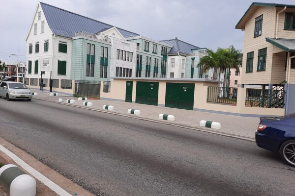 The newer National Assembly building in Paramaribo, a white-and-green government complex seen from the street, with palm trees and a wooden house beside it.