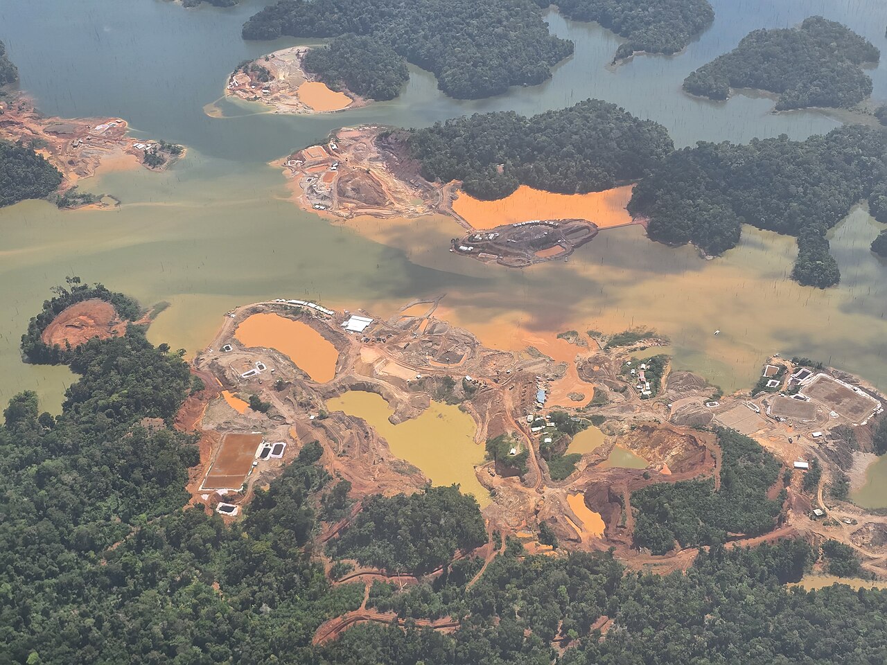 Aerial view of gold-mining sites with orange sediment ponds and cleared red earth on forested islands in a reservoir
