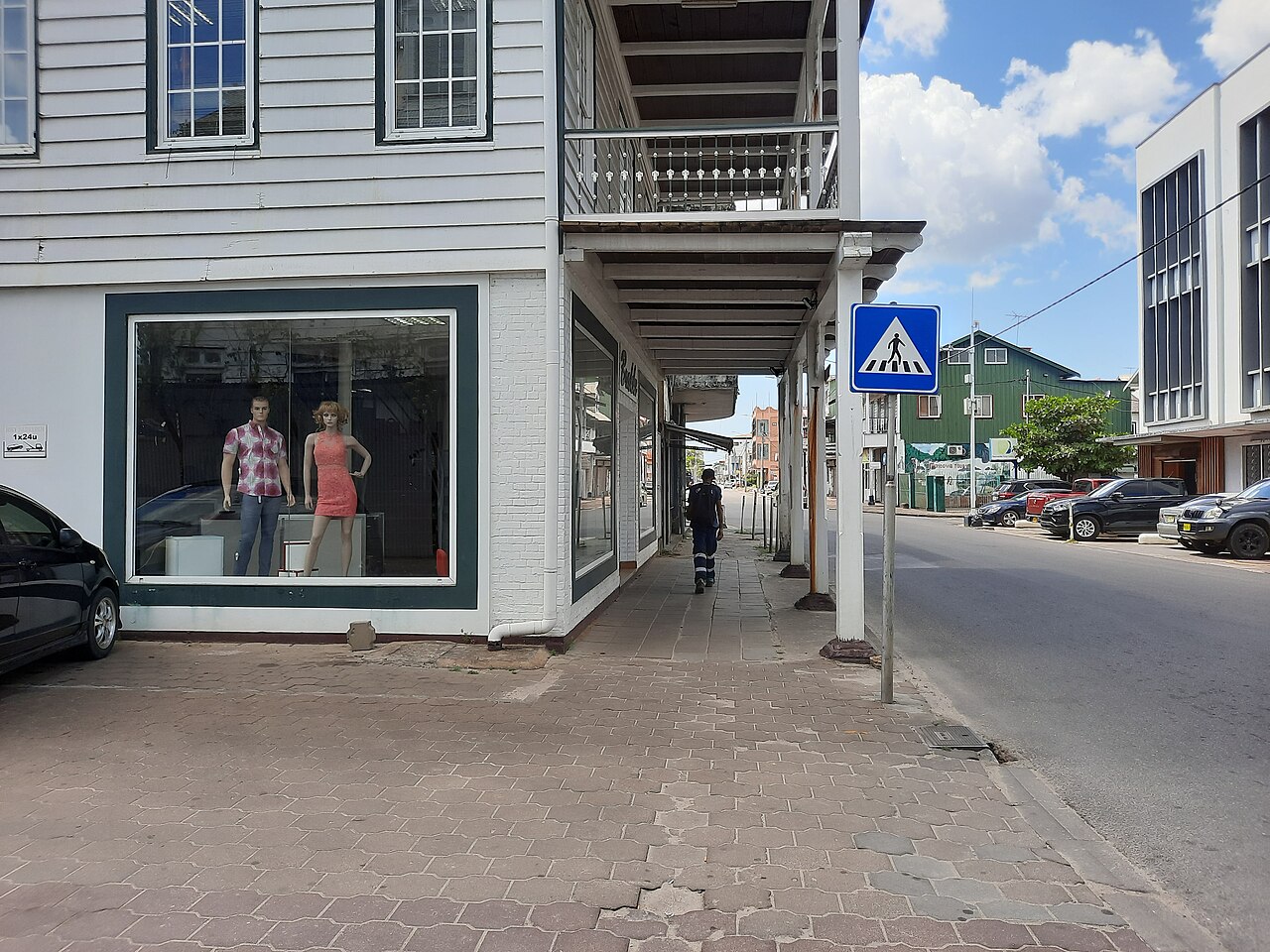 Brick pavement under a wooden arcade on Keizerstraat in Paramaribo, with a pedestrian crossing sign, parked cars and a man walking