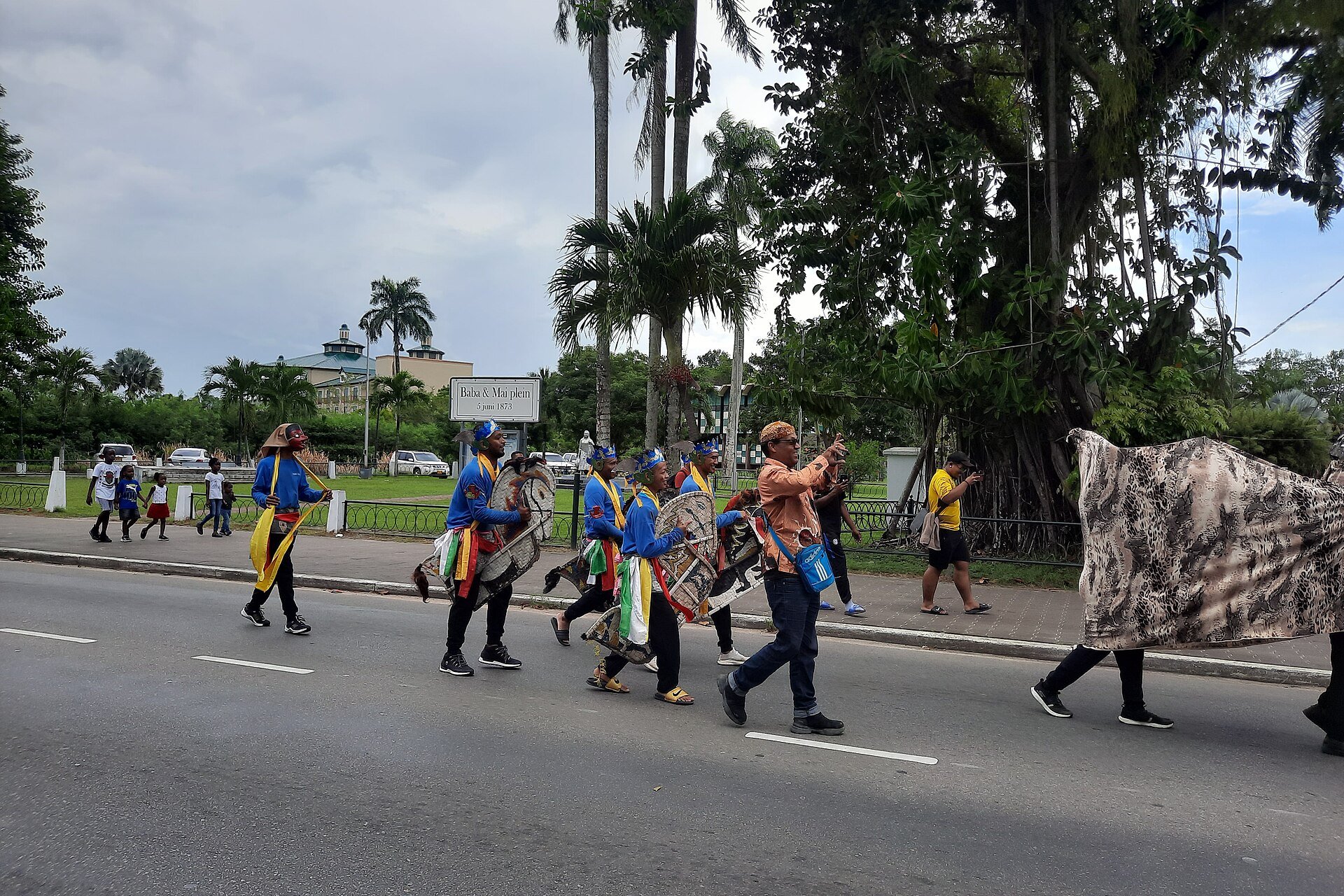 Javanese horse dancers in blue costumes and headcloths carrying woven horse figures along a road in Paramaribo, with a sign reading Baba and Mai plein 5 juni 1873 behind them