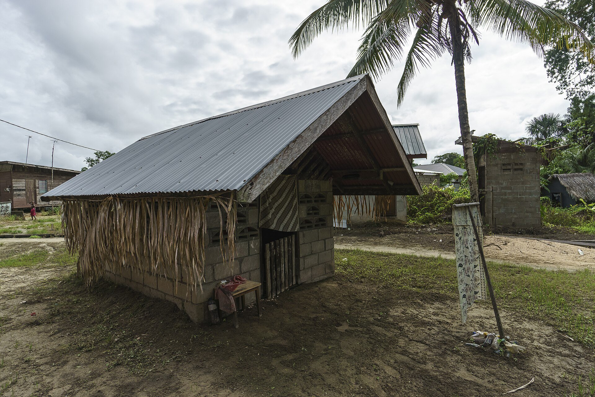 A small concrete-block building with a corrugated metal roof and hanging palm-leaf fringes in a Maroon village, with a coconut palm and wooden houses behind