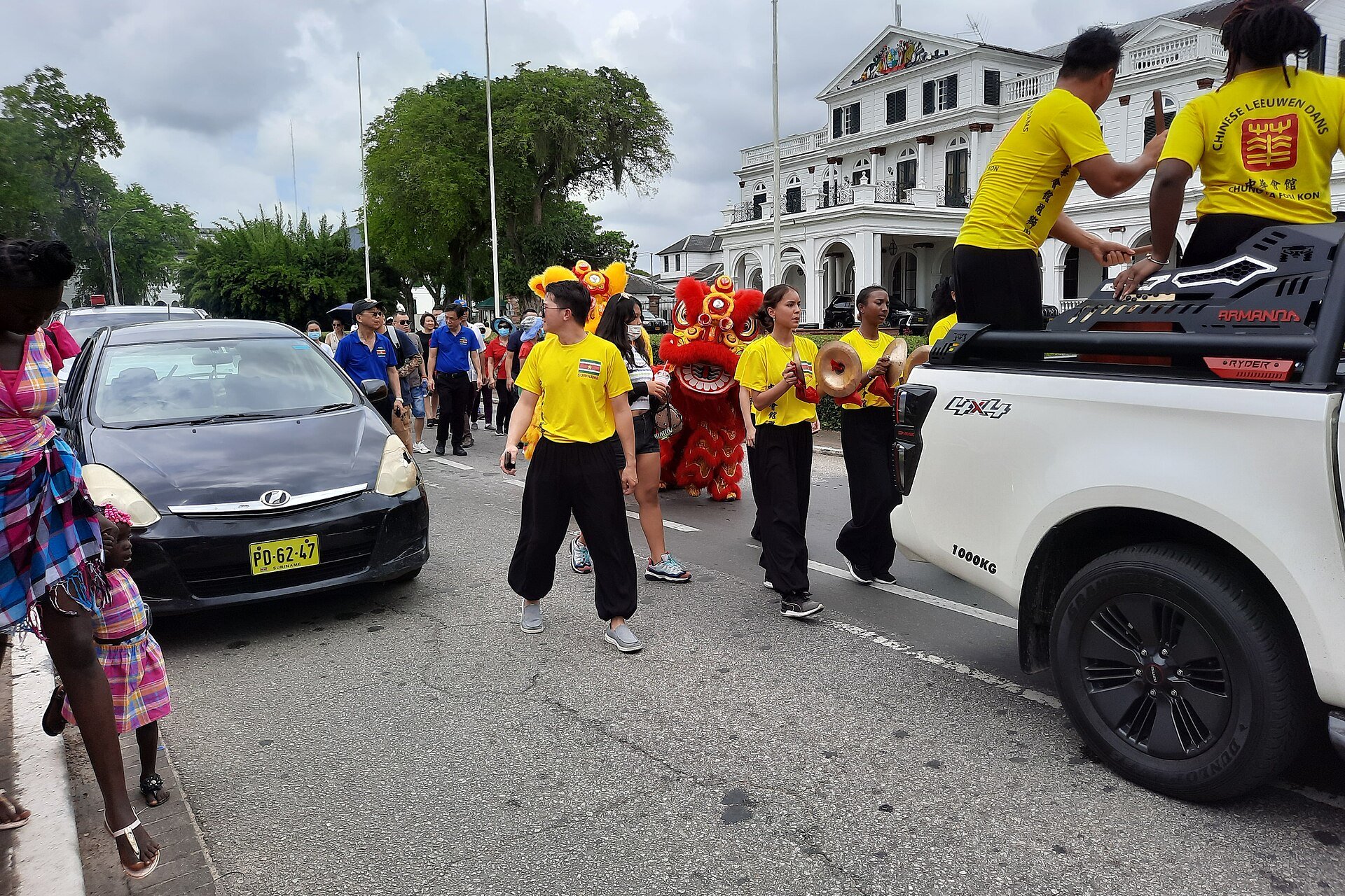 Chinese lion dancers in a red and yellow lion costume walking behind drummers in yellow shirts along a Paramaribo street, with a white colonial building and onlookers