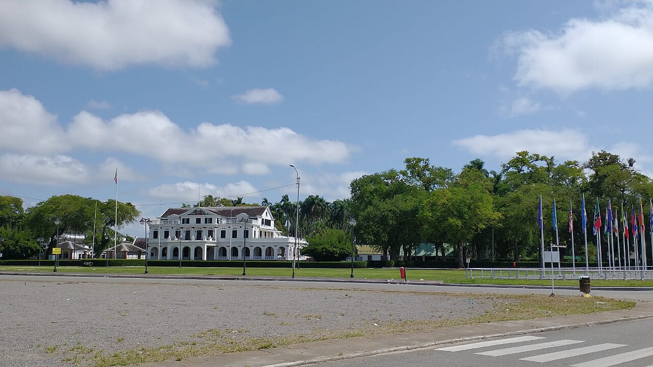 White colonial presidential palace behind a lawn with flagpoles and tall trees in Paramaribo under a blue sky