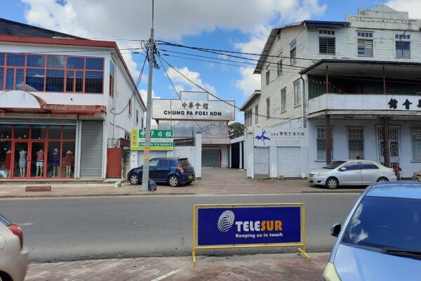 A street in Paramaribo with overhead cables, low commercial buildings and a blue Telesur sign on a barrier in the foreground