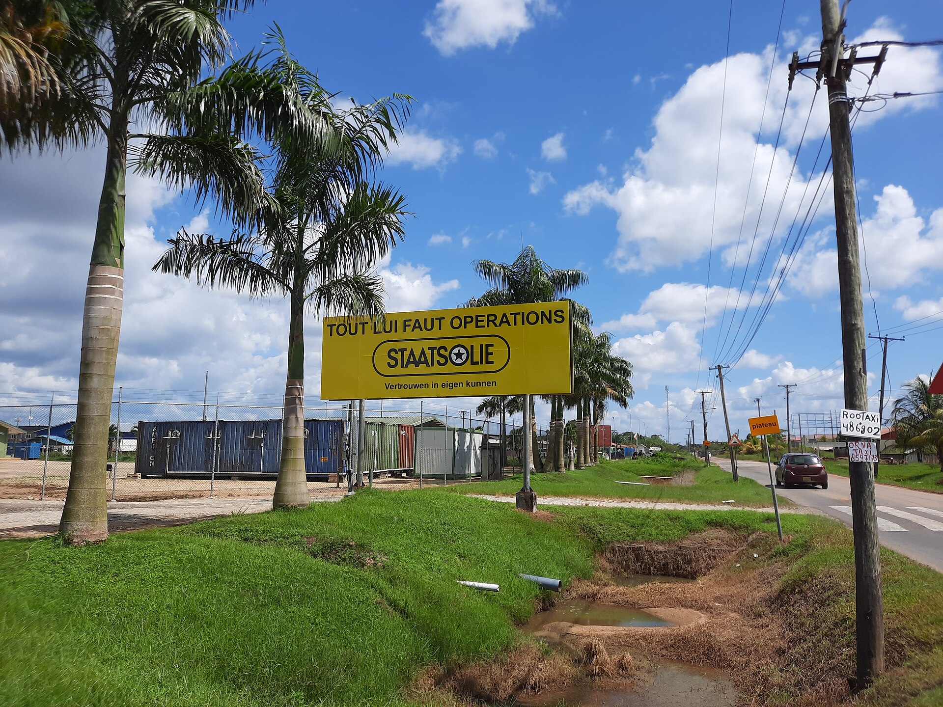 Yellow Staatsolie sign at the Tout Lui Faut operations site beside a road lined with royal palms in Suriname
