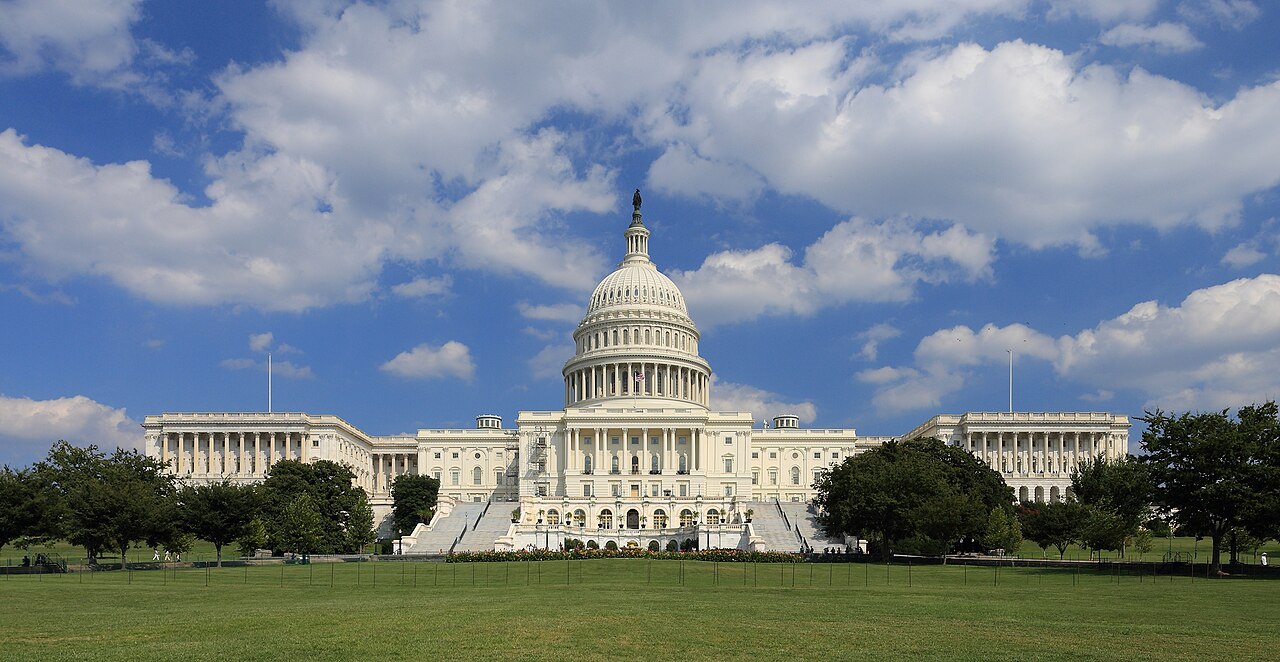 The west front of the US Capitol in Washington, DC, with the dome under a blue sky and a lawn in front
